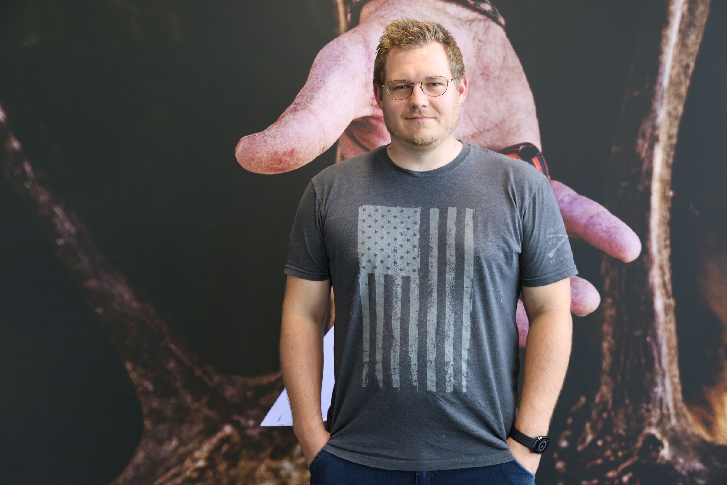 Colten Krauter in glasses and a gray T-shirt, in front of a large print of a weathered hand